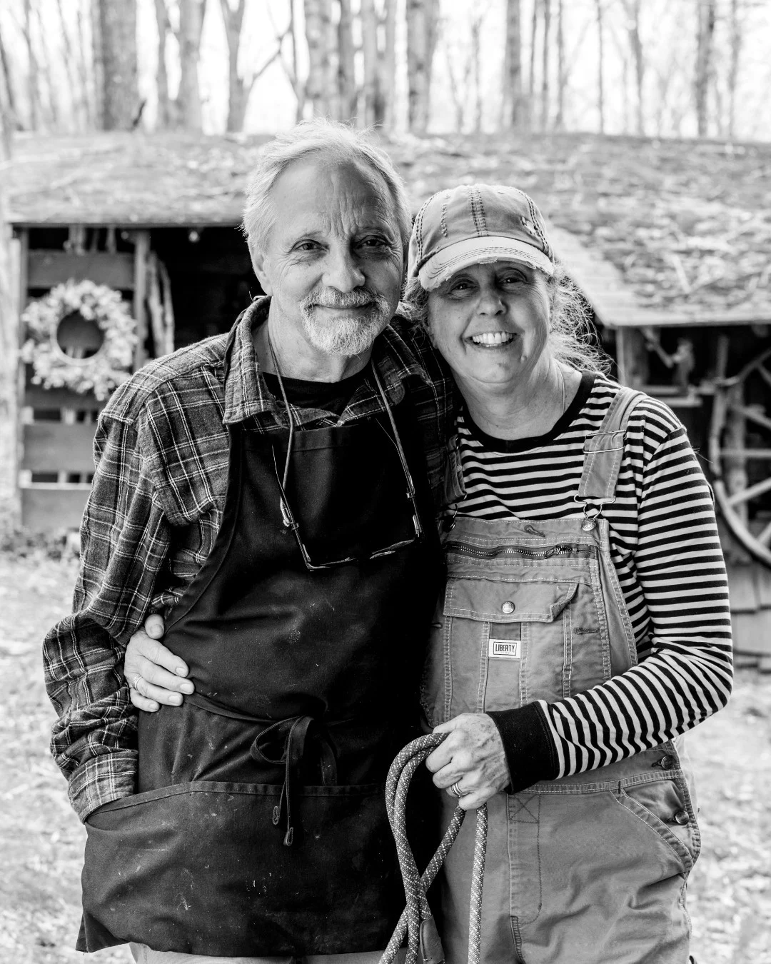 A smiling couple in work clothes stands arm in arm outside a wooden barn.