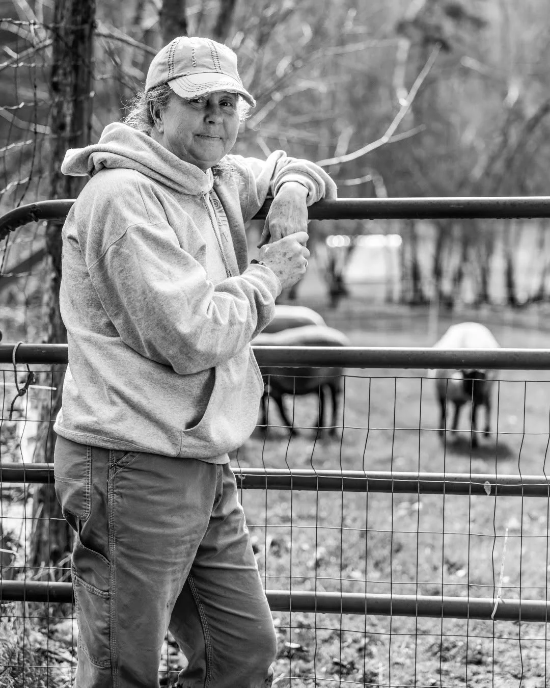 A woman rests her arms on a livestock gate with sheep behind her.