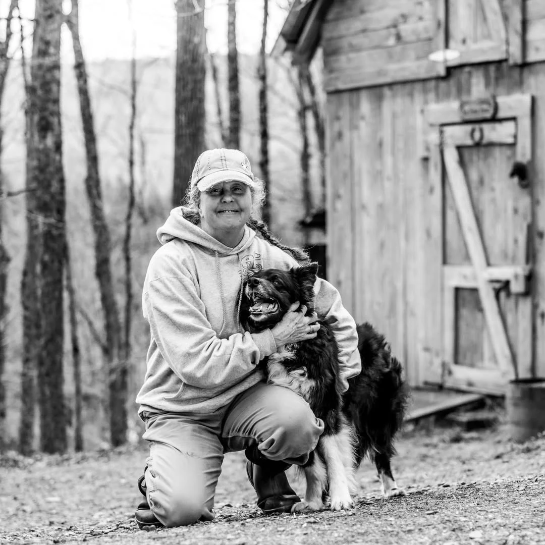A woman kneels beside a black-and-white dog outside a wooden barn.