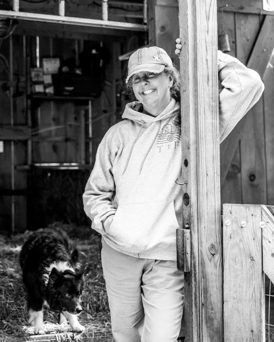 A smiling woman leans against a barn post with a dog behind her.