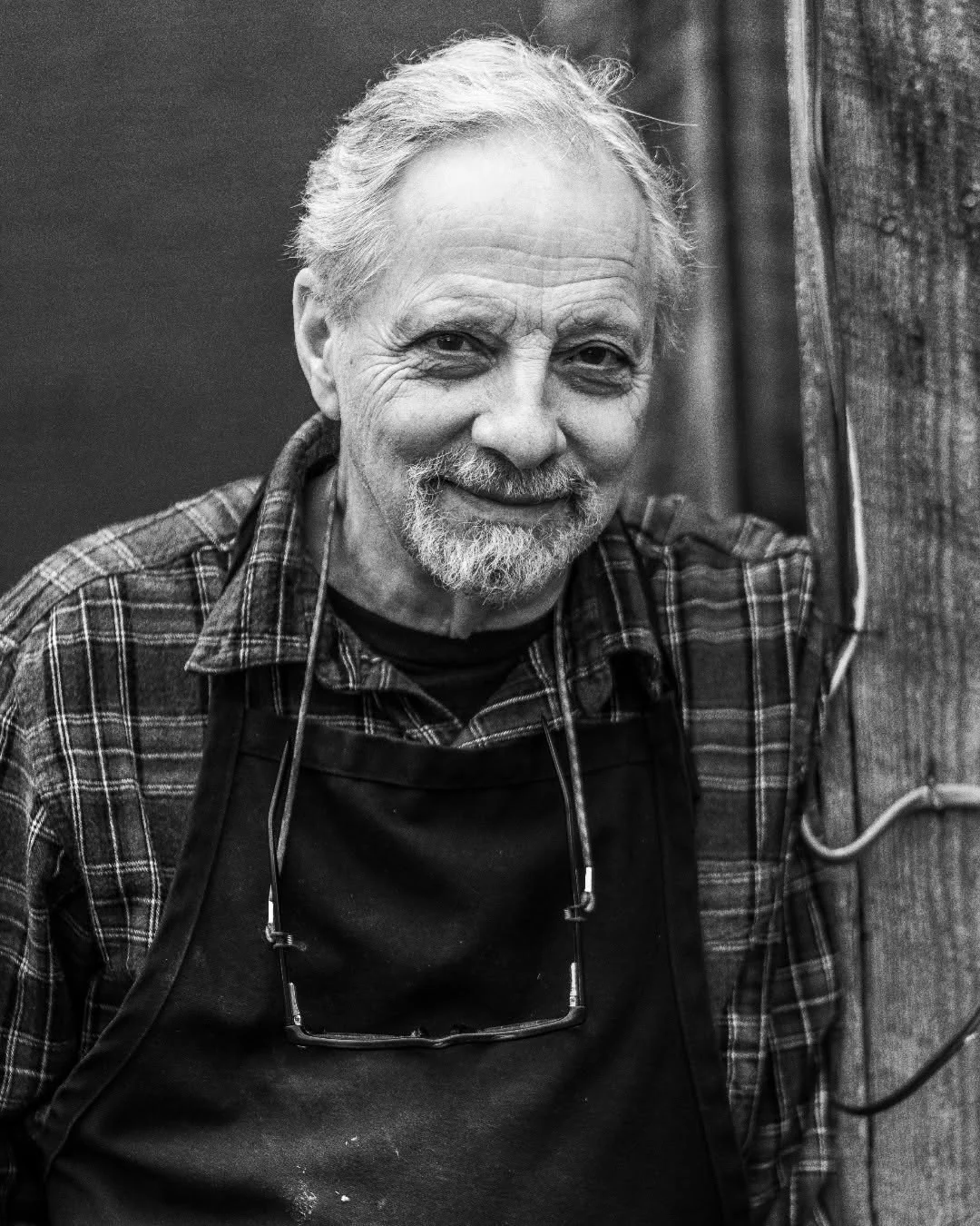 A smiling man wearing a plaid shirt stands beside a barn post.