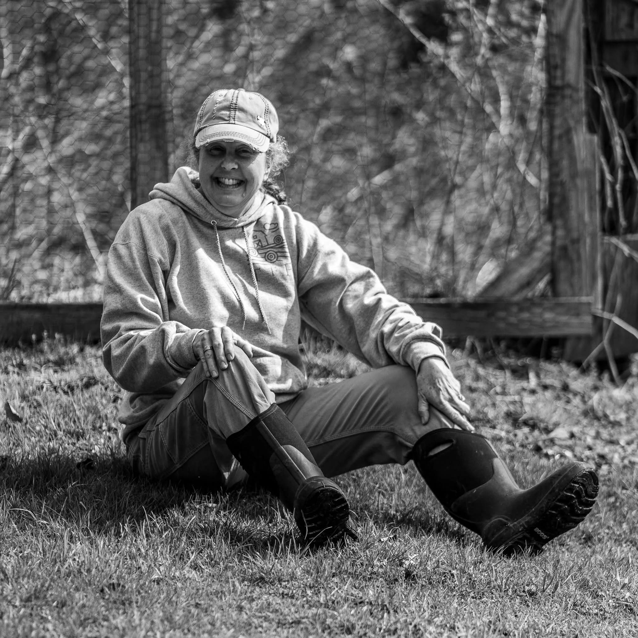 A woman sits outdoors beside a barn with her hands folded in her lap.