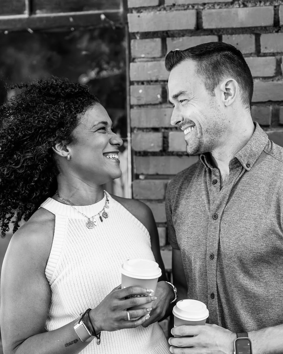 A couple holding paper cups smiles at each other against a brick wall.