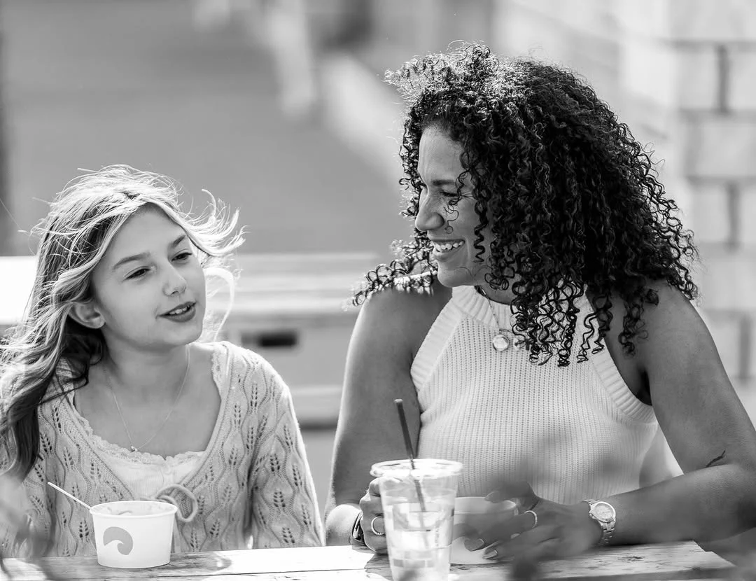 A woman talks with a young girl at an outdoor cafe table.