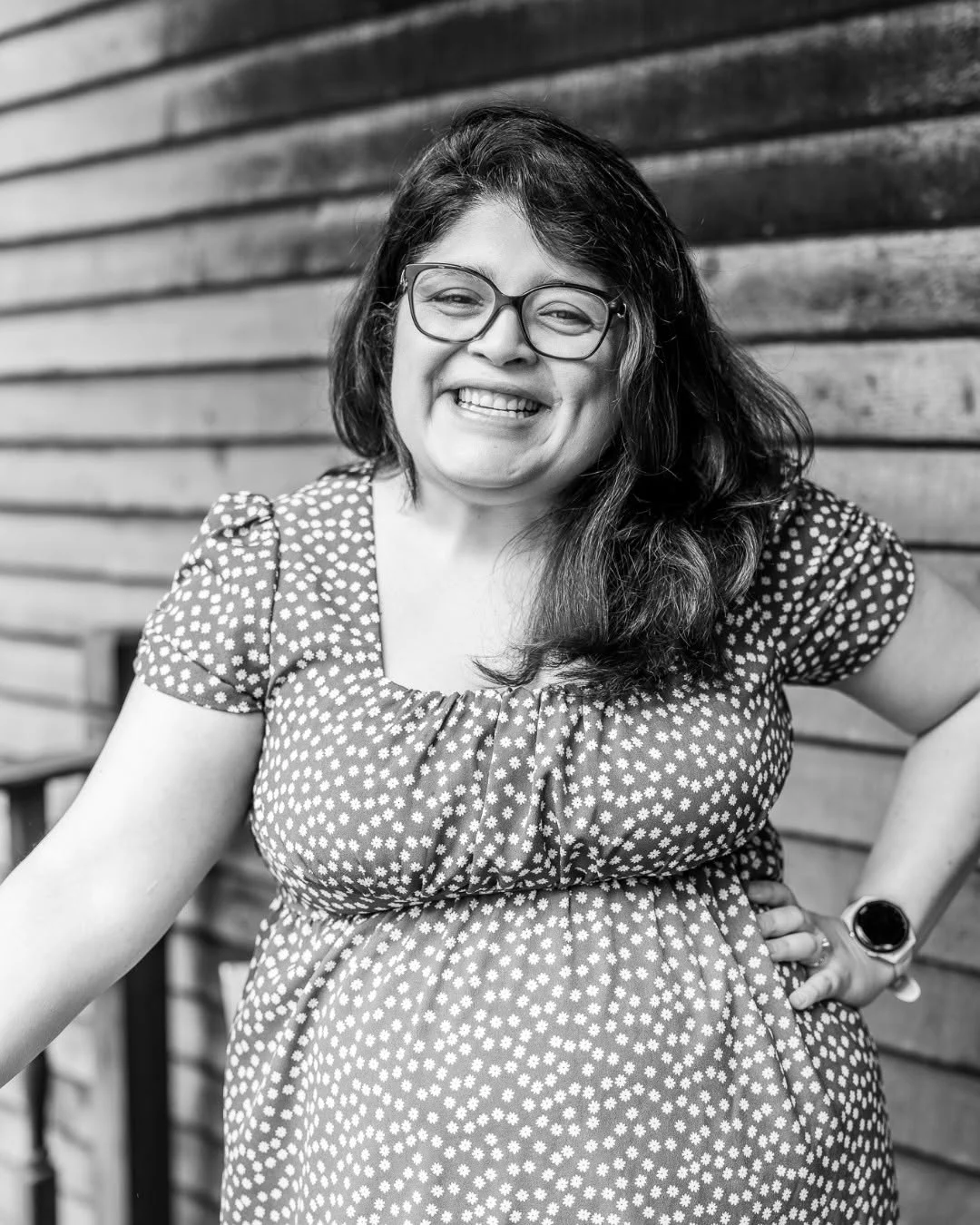 A smiling woman sits against the wooden siding of a porch.