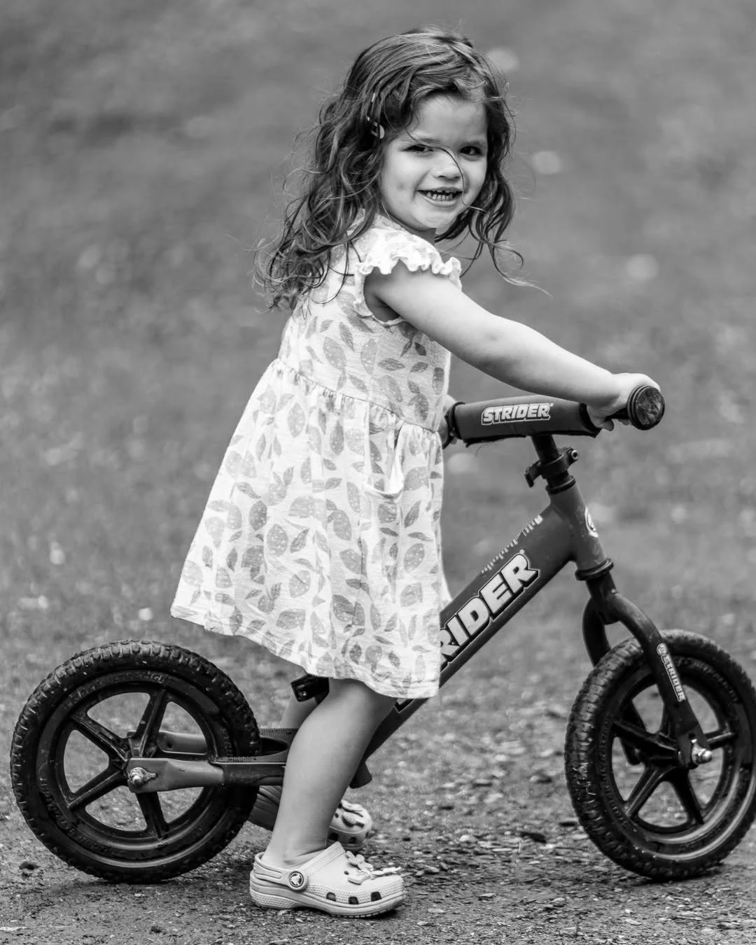 A young girl smiles while riding a balance bike on grass.