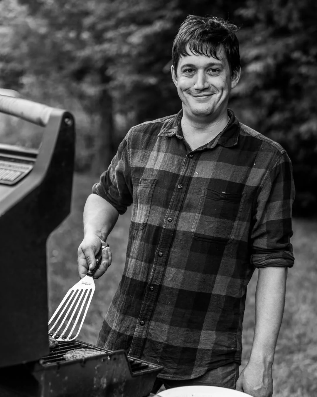 A smiling man holds a spatula beside an outdoor grill.