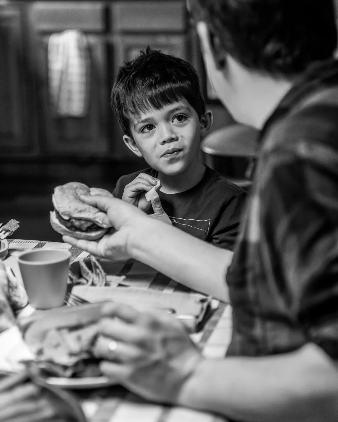 A boy looks across a dining table while an adult holds a sandwich.