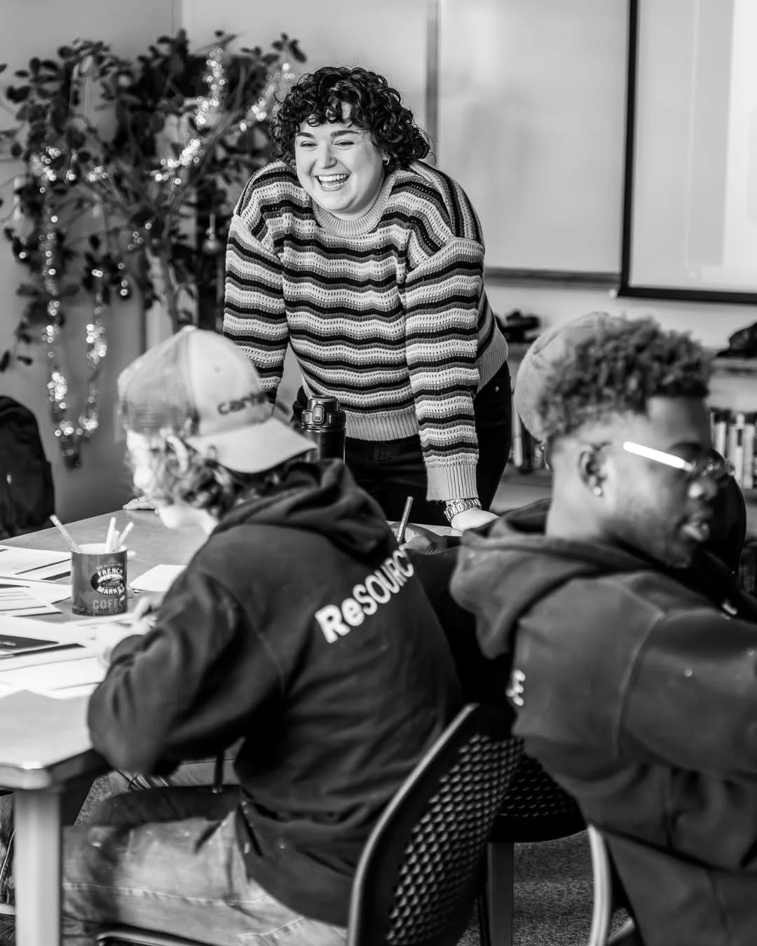 A smiling instructor stands beside ReSOURCE participants seated at a table.