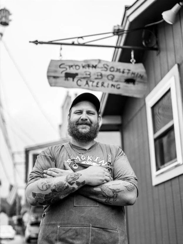 A bearded man in a cap and apron stands with folded arms outside Smokin’ Somethin’ BBQ.