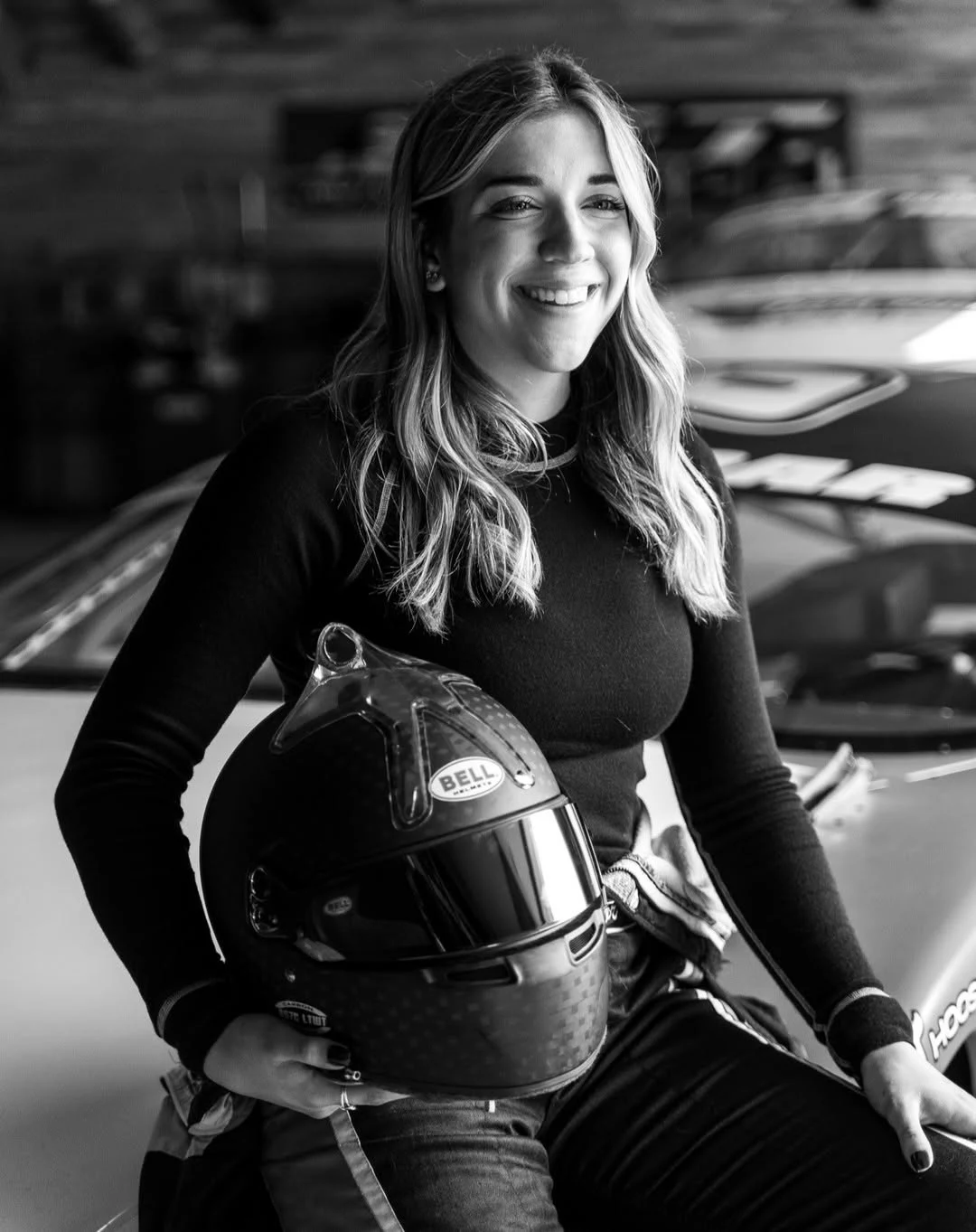 Black-and-white portrait of Taylor Hoar smiling with her helmet beside a stock car.