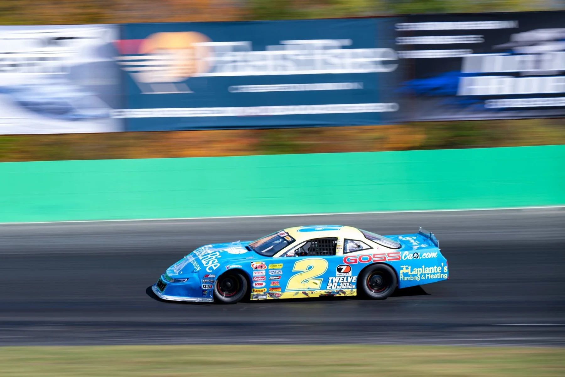 A blue No. 12 stock car streaks past the green retaining wall with the background blurred.