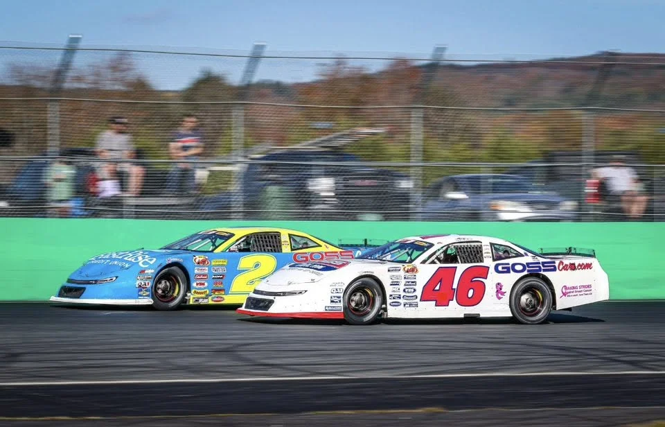A blue No. 2 car and a white No. 46 car race side by side past spectators lining the fence at Thunder Road.