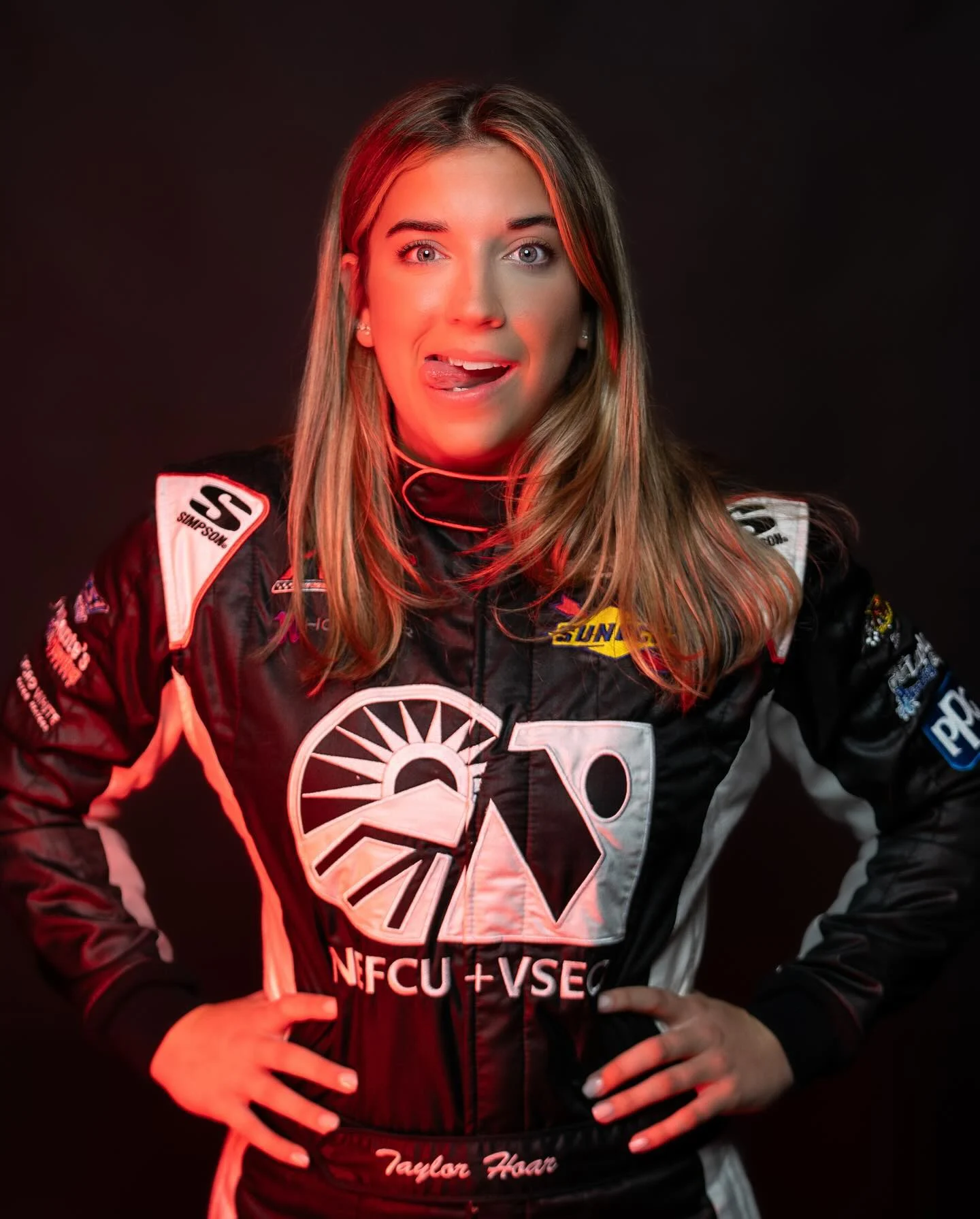 Taylor Hoar sticks out her tongue in a red-lit portrait wearing her black racing suit.