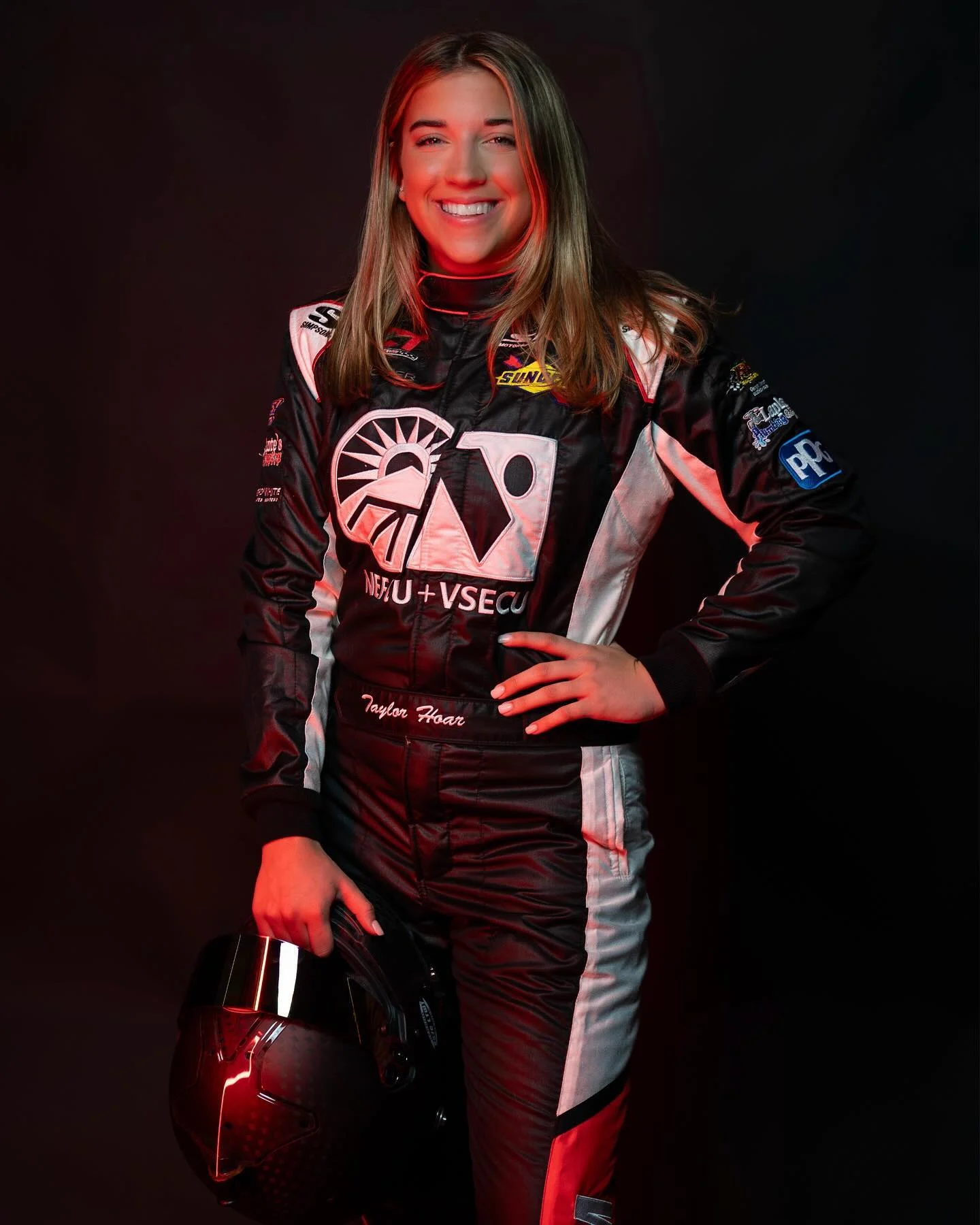 Taylor Hoar smiles in a full-length portrait, holding her helmet under red studio light.