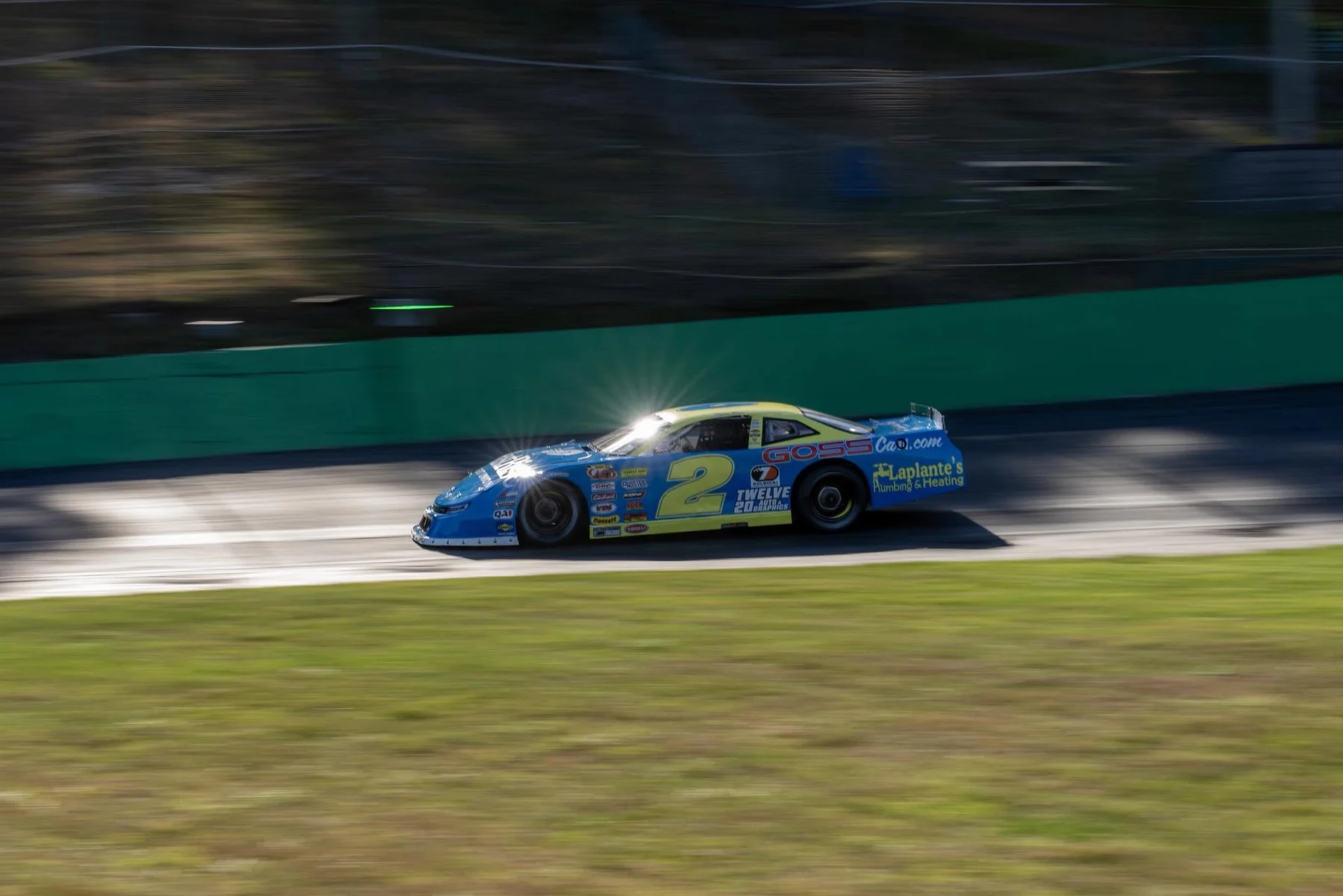 A blue and yellow No. 2 stock car streaks past the green retaining wall, sunlight glinting off its roof.