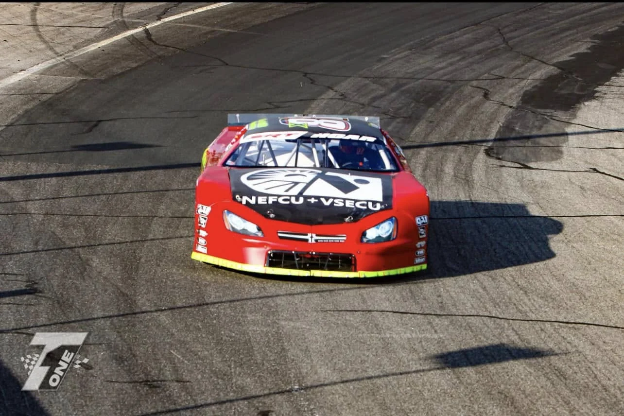 A red stock car with a NEFCU + VSECU hood logo rounds a banked turn.