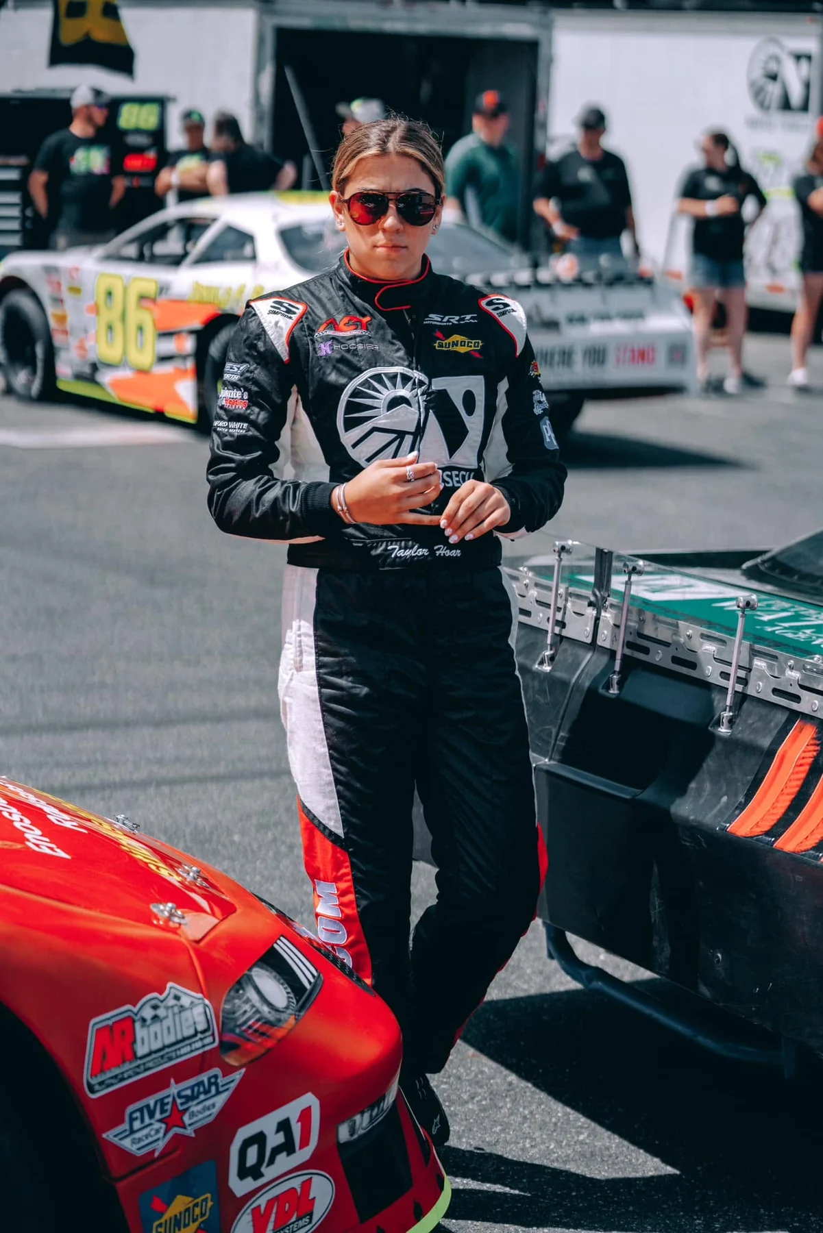 Taylor Hoar in sunglasses and a black race suit stands between stock cars in the Thunder Road pit area.