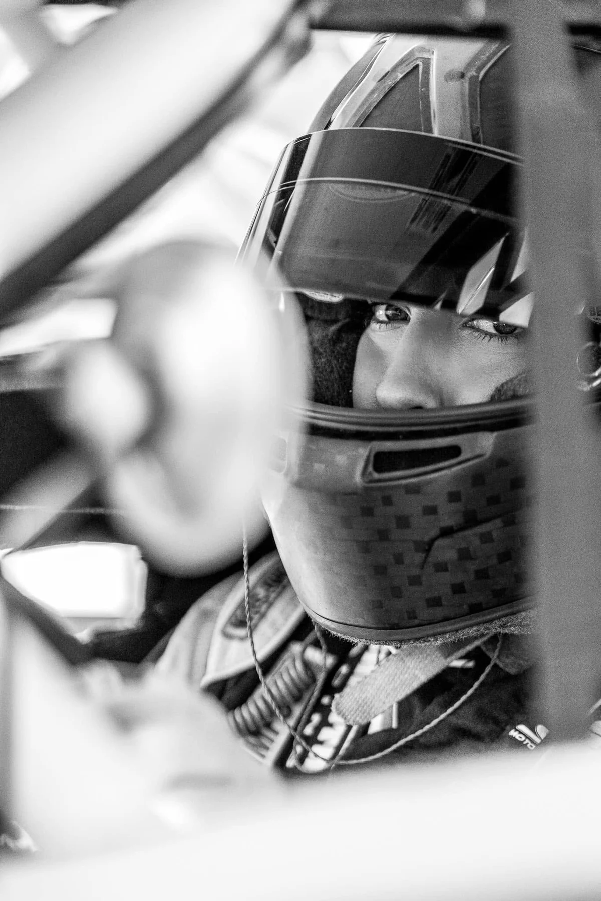 Black-and-white view through a stock car's roll cage of a driver in a full-face helmet waiting in the cockpit.