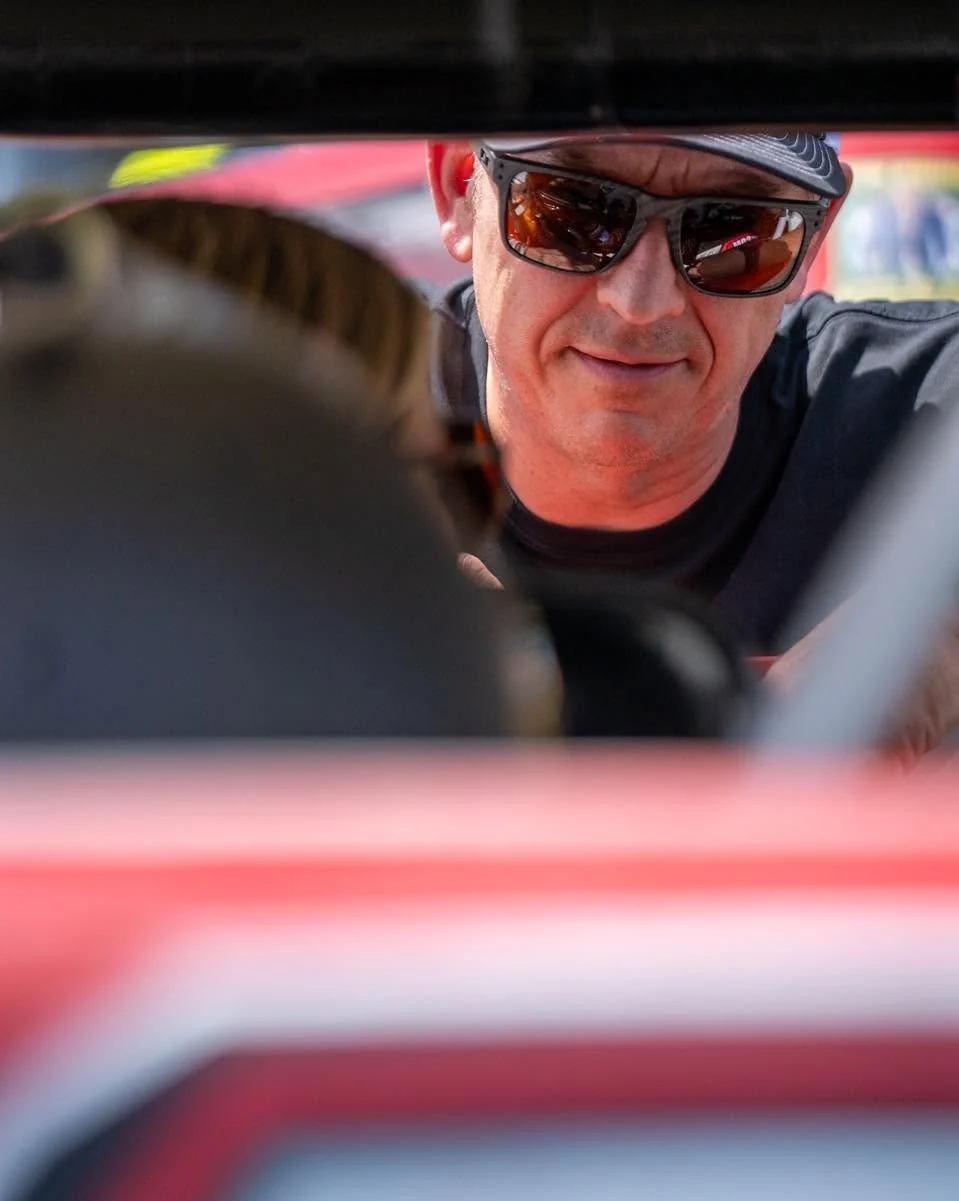 A crew member in sunglasses looks through the open window of a red stock car.