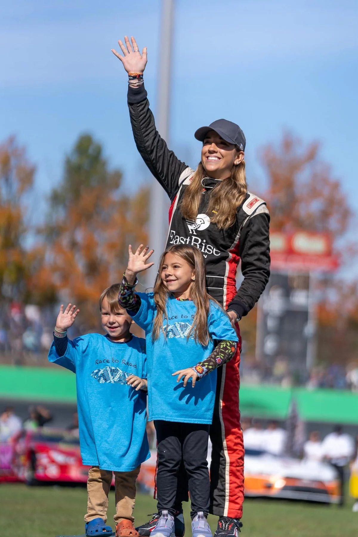 Taylor Hoar in an EastRise race suit waves to the crowd beside two children in blue Race to Read T-shirts at Thunder Road.