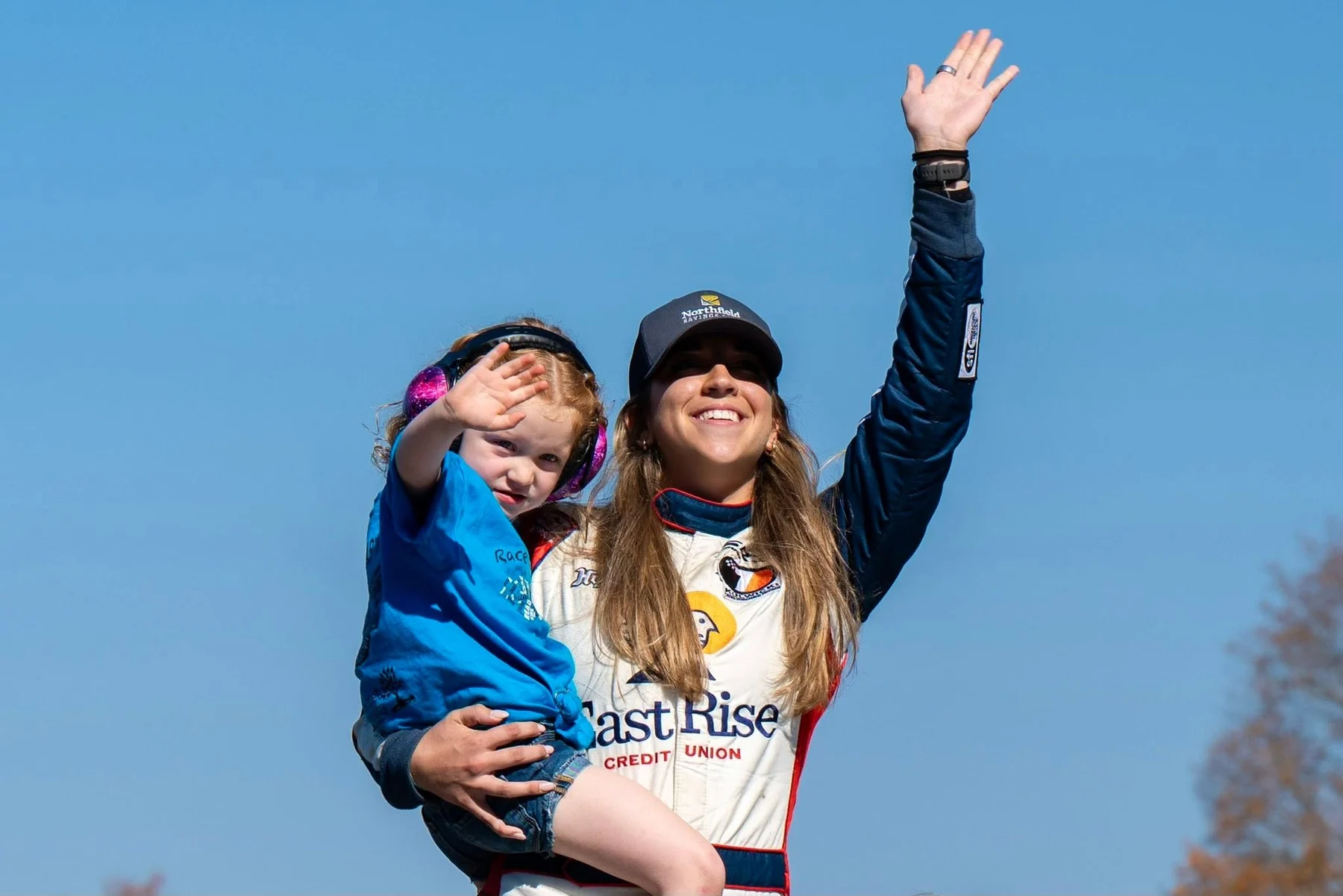Taylor Hoar in an EastRise Credit Union race suit waves while holding a young child wearing ear protection under a blue sky.