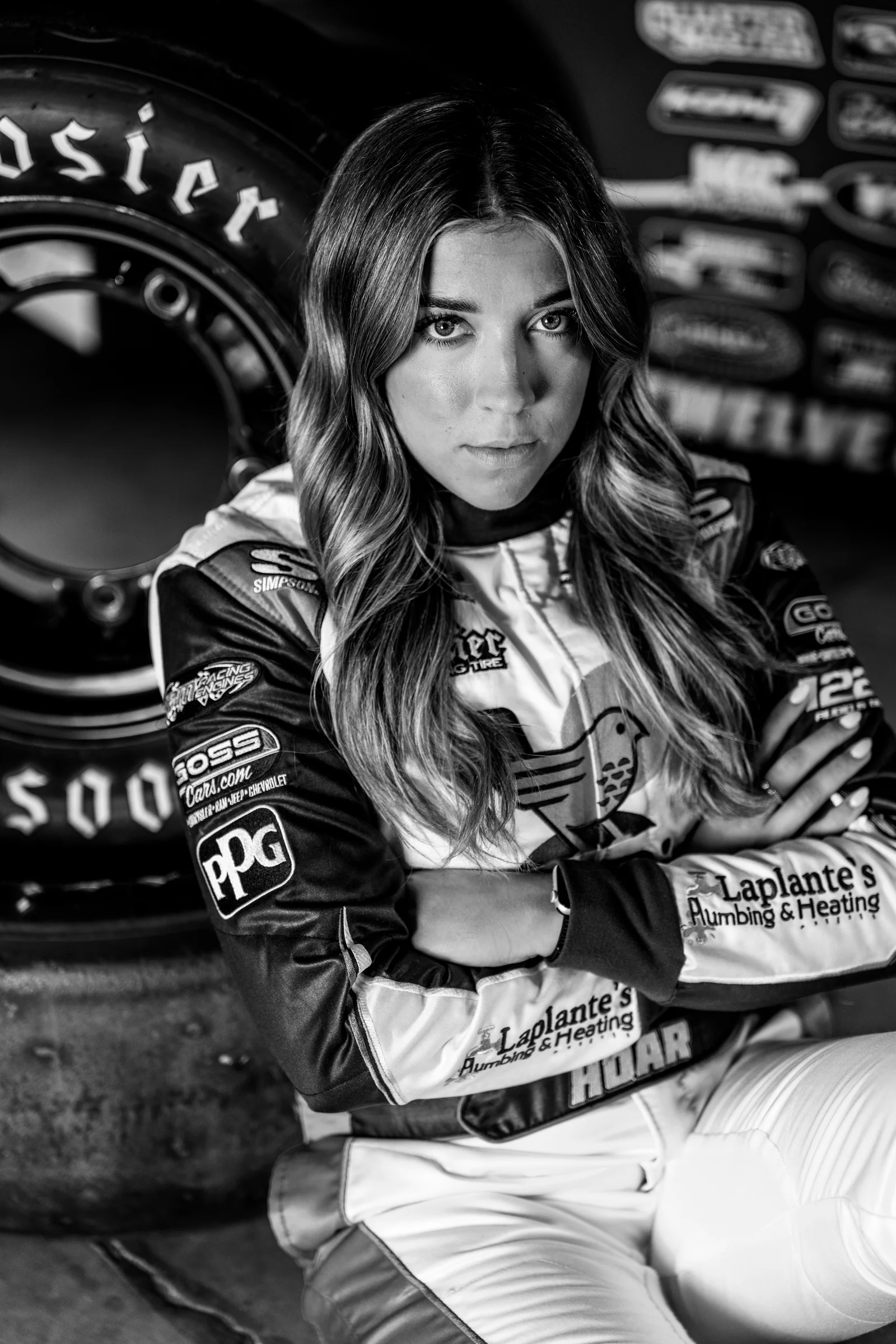 Black-and-white portrait of Taylor Hoar seated in her race suit inside the race shop.