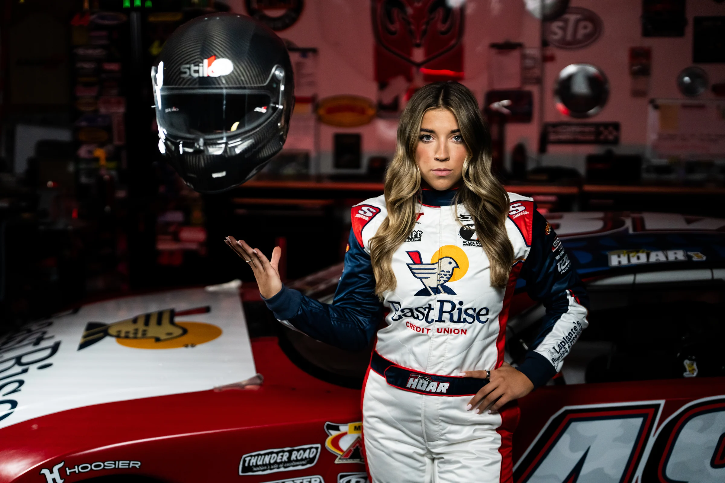 Taylor Hoar stands beside the No. 48 car in her EastRise race suit, holding her helmet at shoulder height.