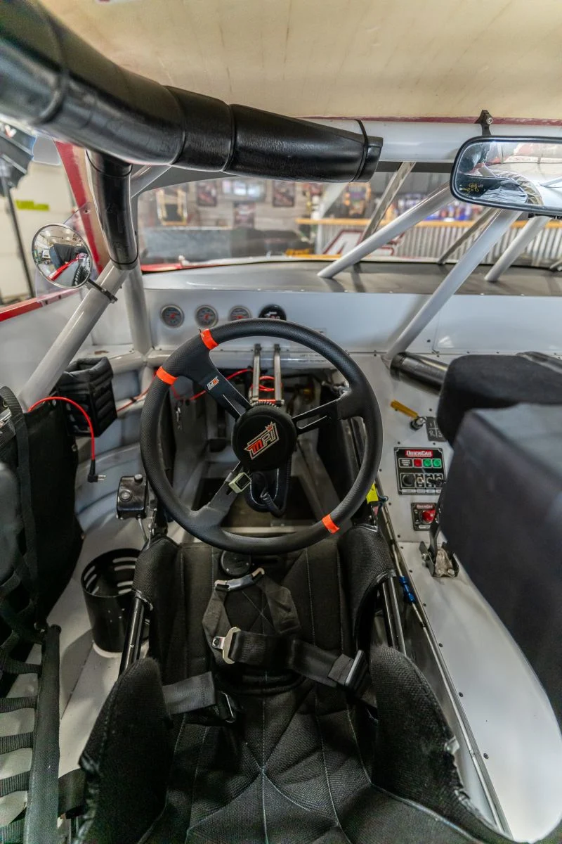 Interior of a stock race car in a garage, showing a padded roll cage, removable steering wheel, gauges, and a racing seat with harness.
