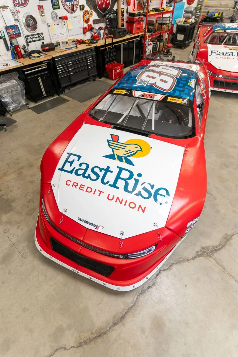 Taylor Hoar's No. 48x race car with a large EastRise Credit Union hood logo parked in the race shop, a matching car behind it.