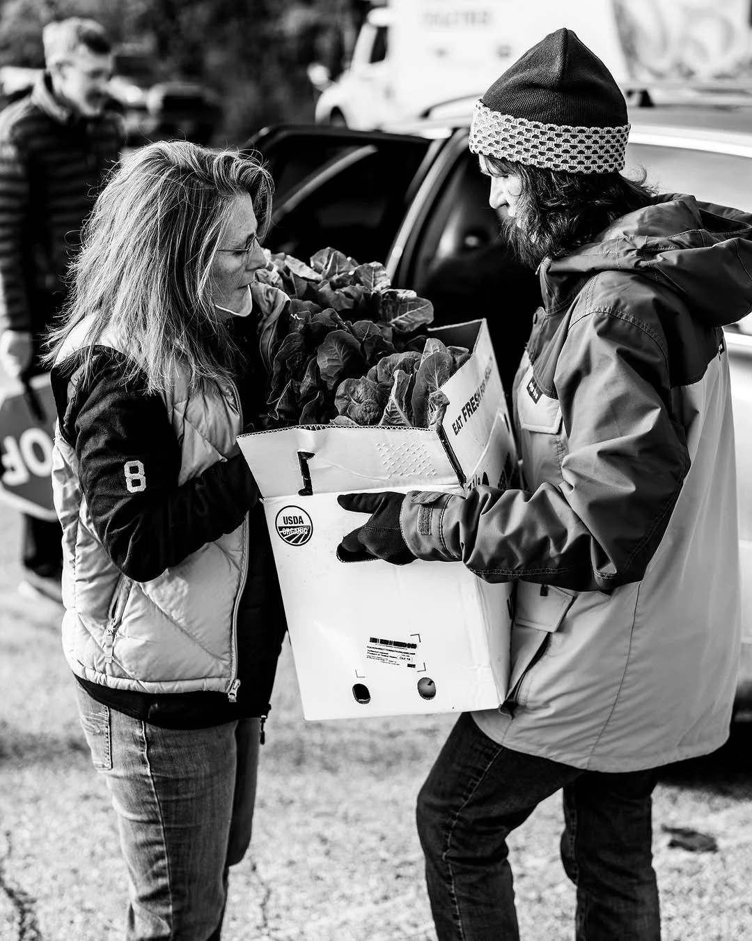 Two volunteers transfer a box of produce beside a car.