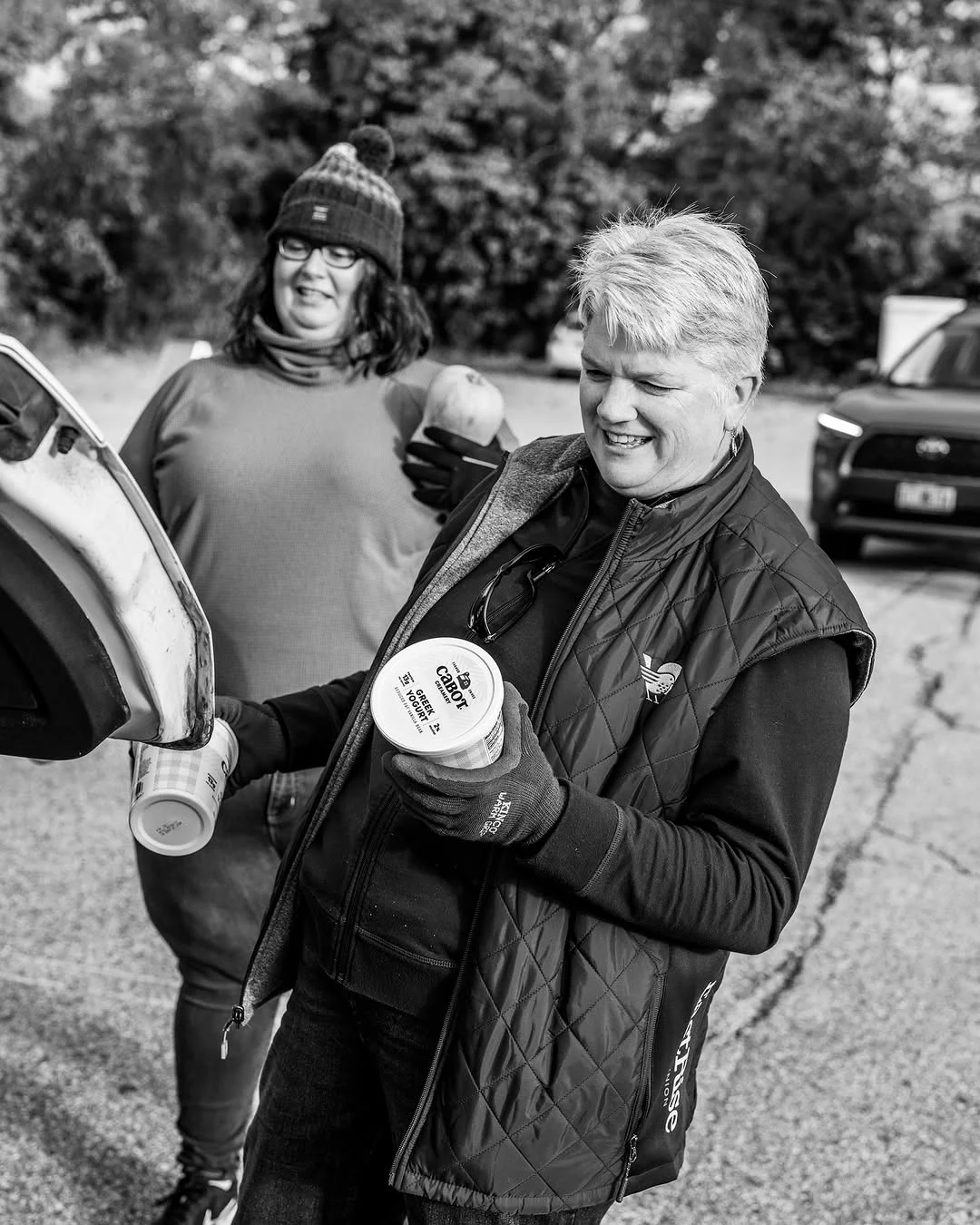 Two volunteers carry tubs of yogurt toward an open car.