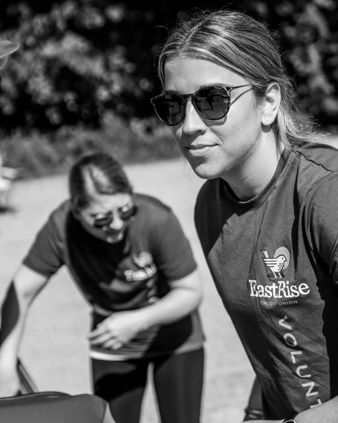 A volunteer wearing sunglasses stands beside boxes while another packs produce.