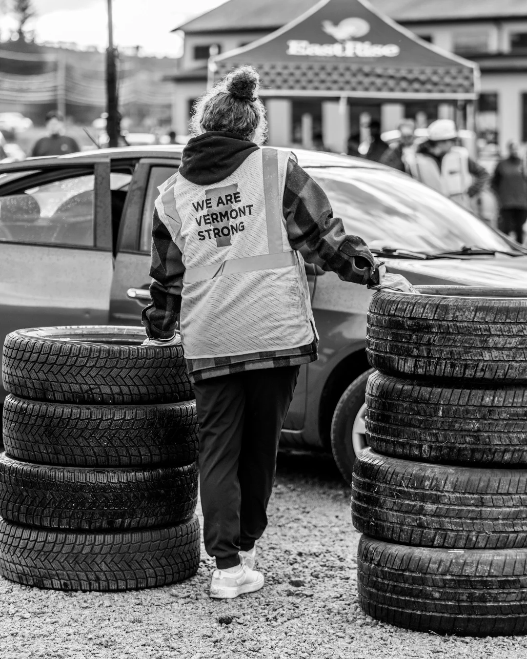 A volunteer stands among stacked tires beside a parked car.