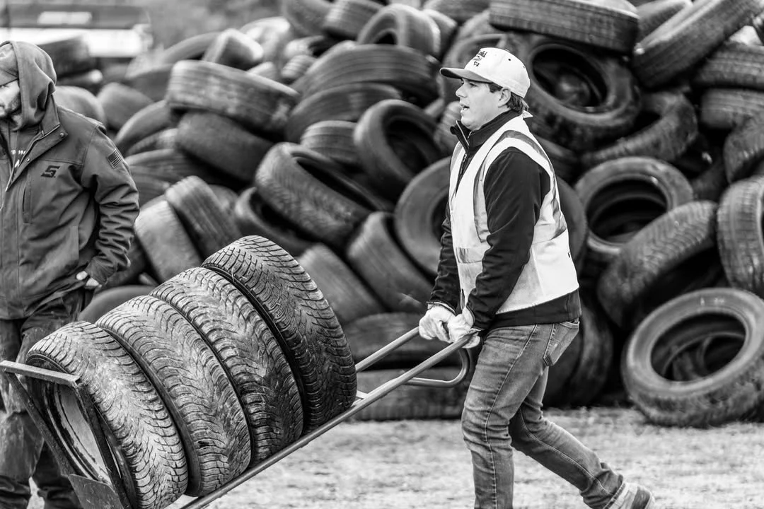 A volunteer pushes four tires on a dolly past a towering tire pile.