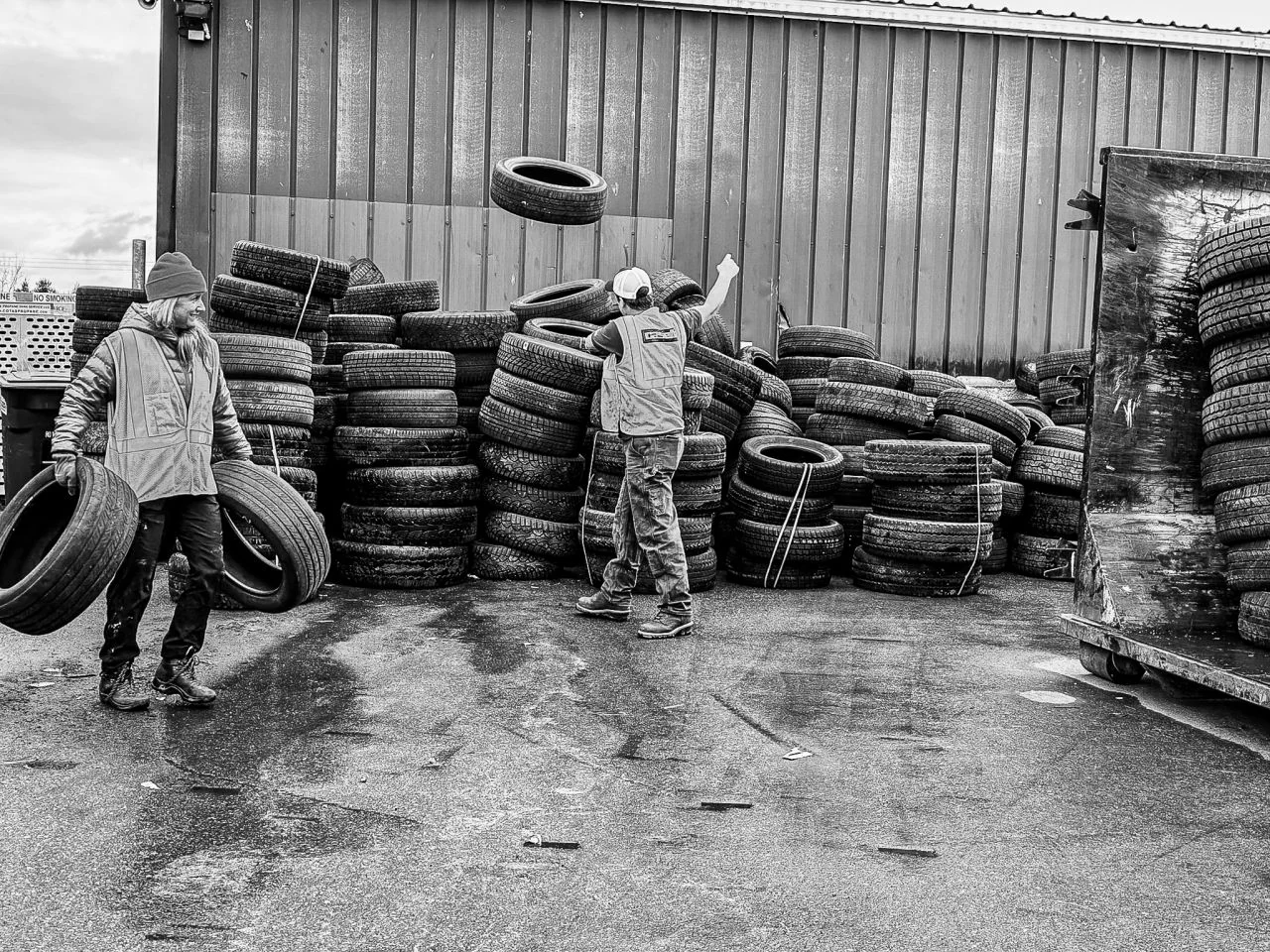 Volunteers stack tires beside a warehouse during a rainy collection day.