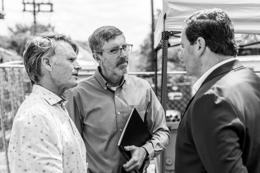 Three men talk near a canopy while one holds a black binder.