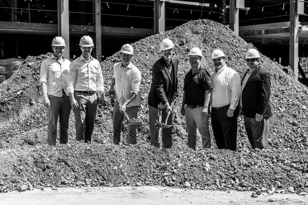 A group in hard hats poses with shovels beside a dirt mound.