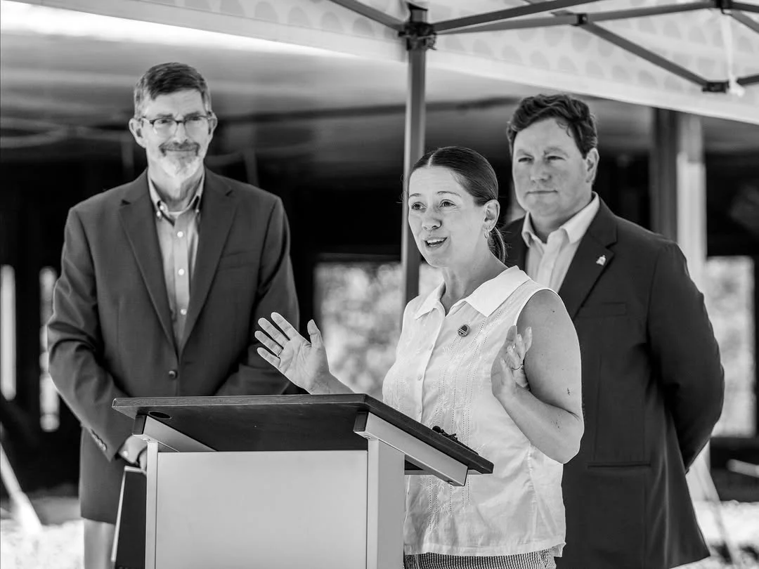 A woman gestures while speaking at a lectern under a tent, with two men behind her.