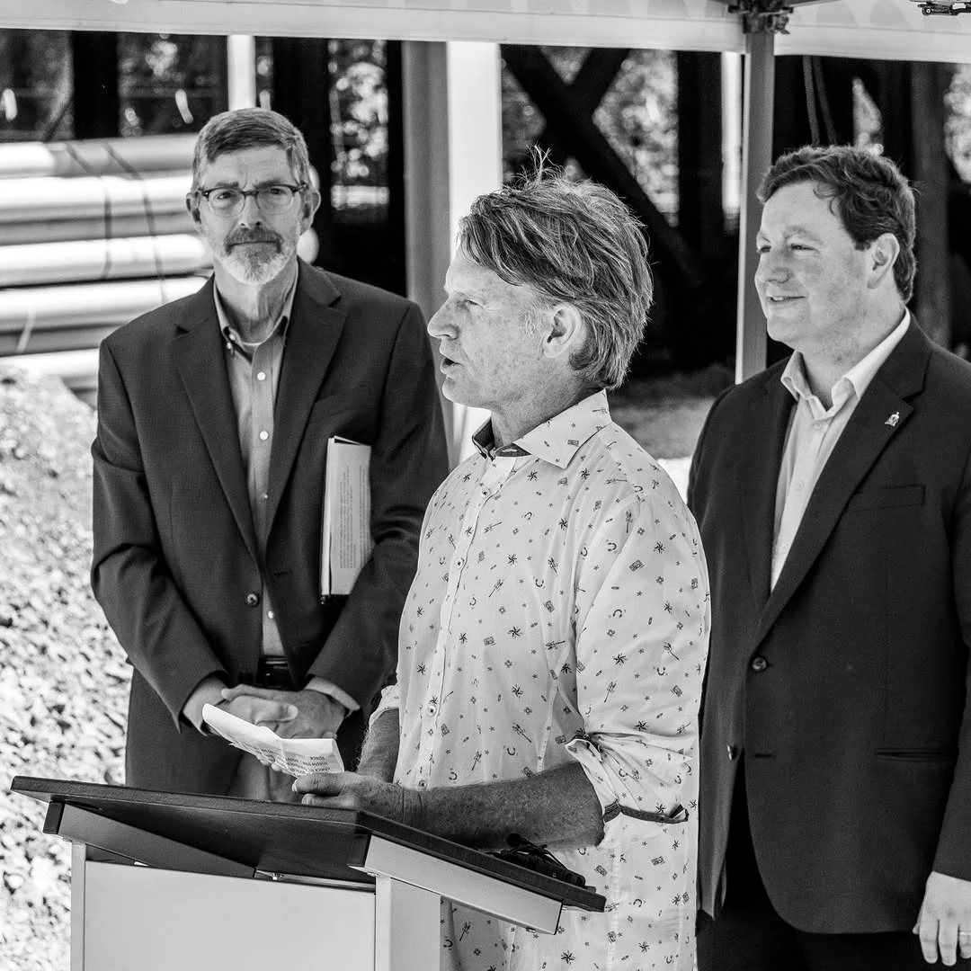 A woman in a patterned blouse speaks at a lectern while two men listen.