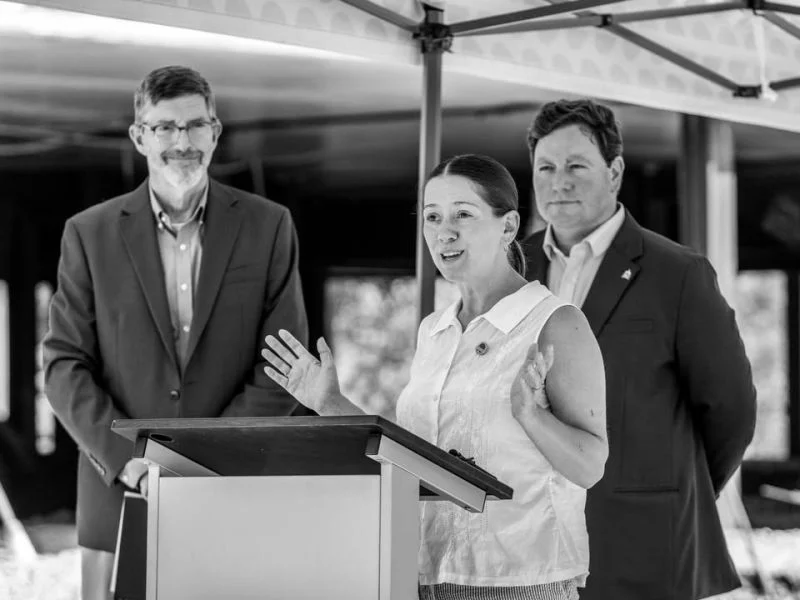 A woman gestures while speaking at a lectern under a tent, with two men behind her.