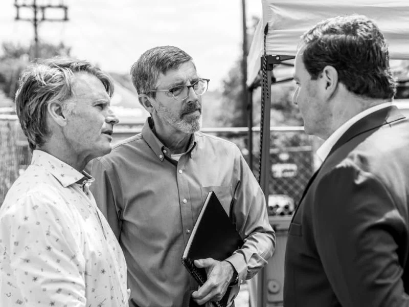 Three men talk near a canopy while one holds a black binder.
