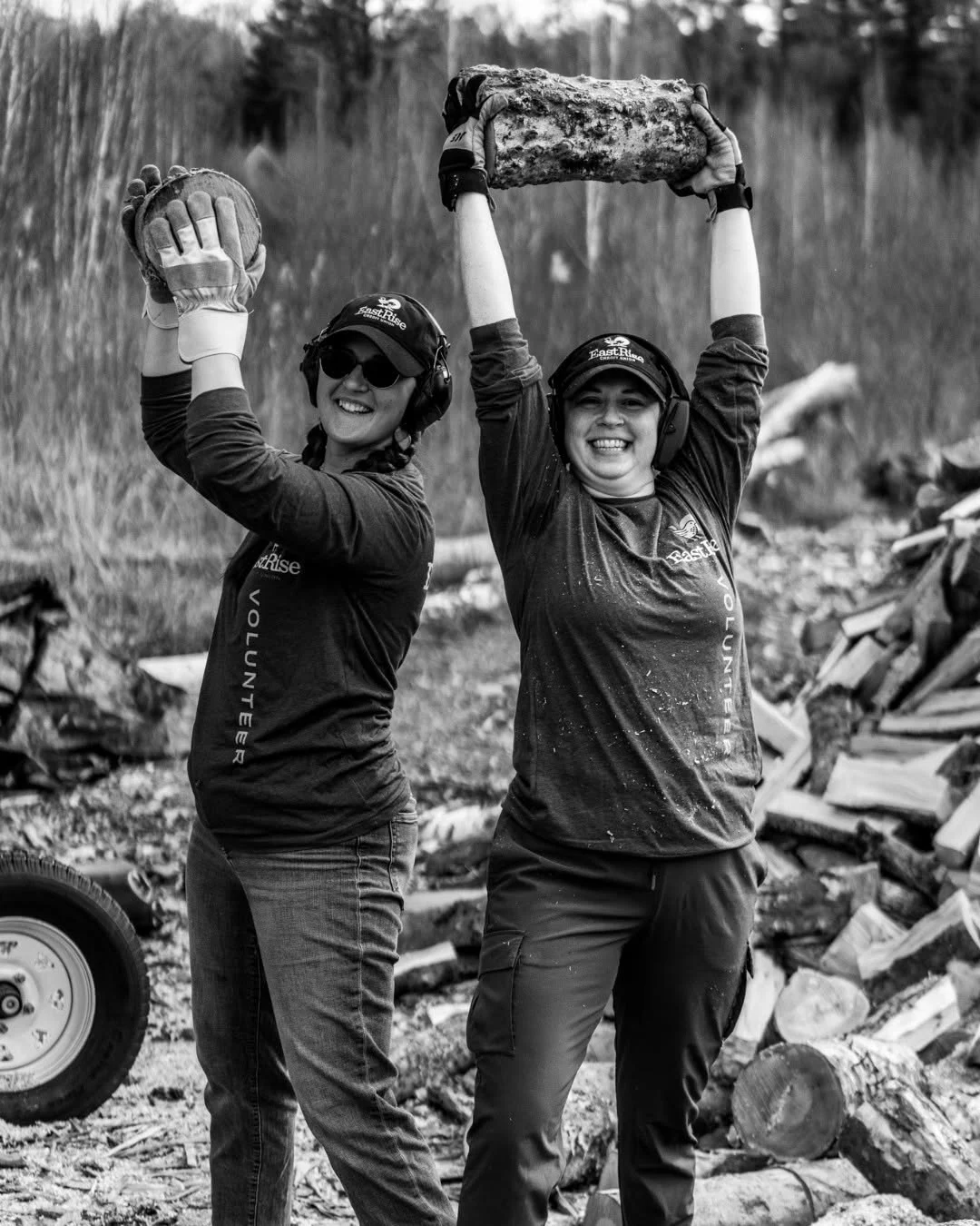 Two smiling volunteers lift logs overhead beside a woodpile.