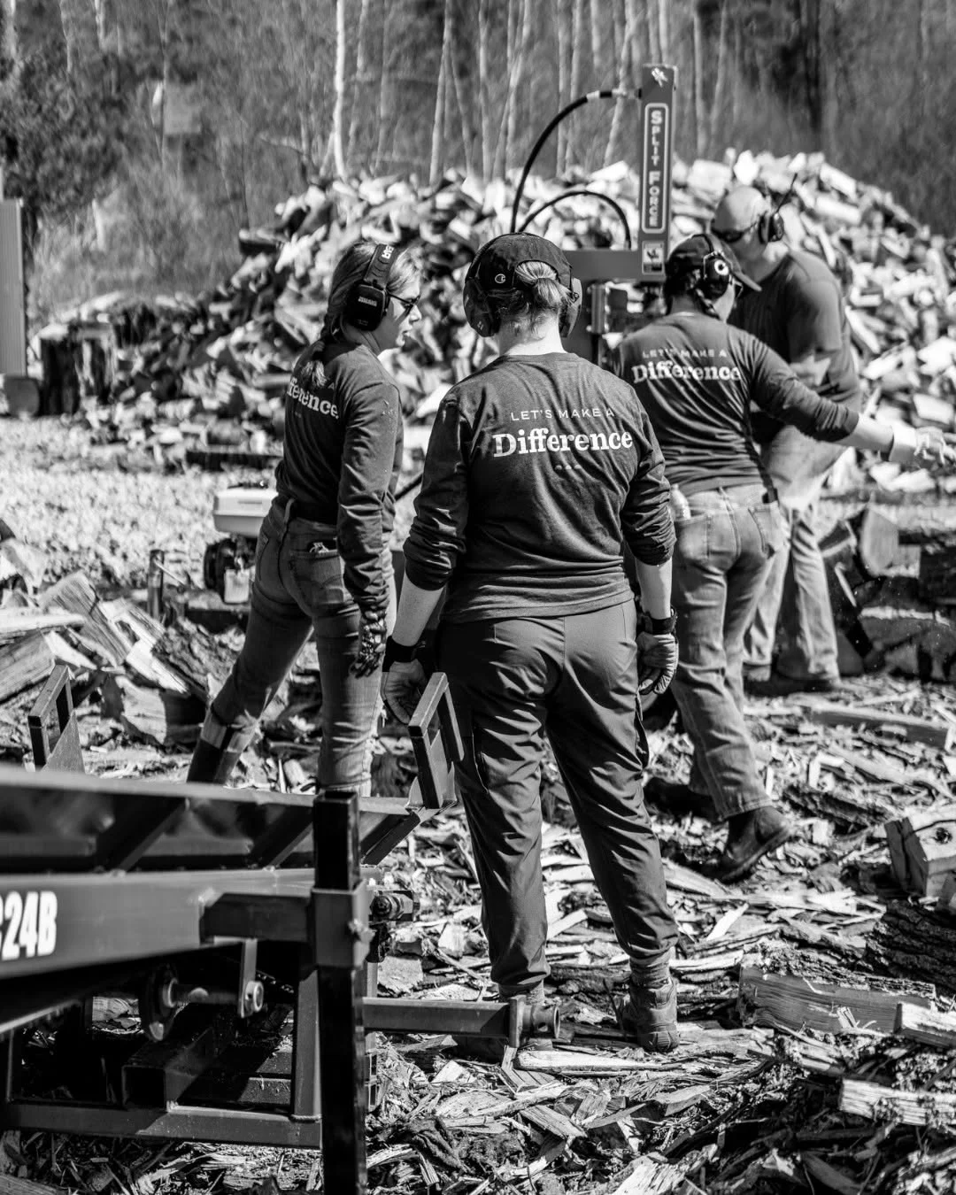 Volunteers feed logs into a processing machine beside a large woodpile.