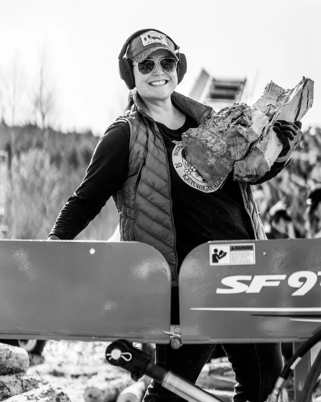 A smiling volunteer carries an armful of split firewood past a machine.