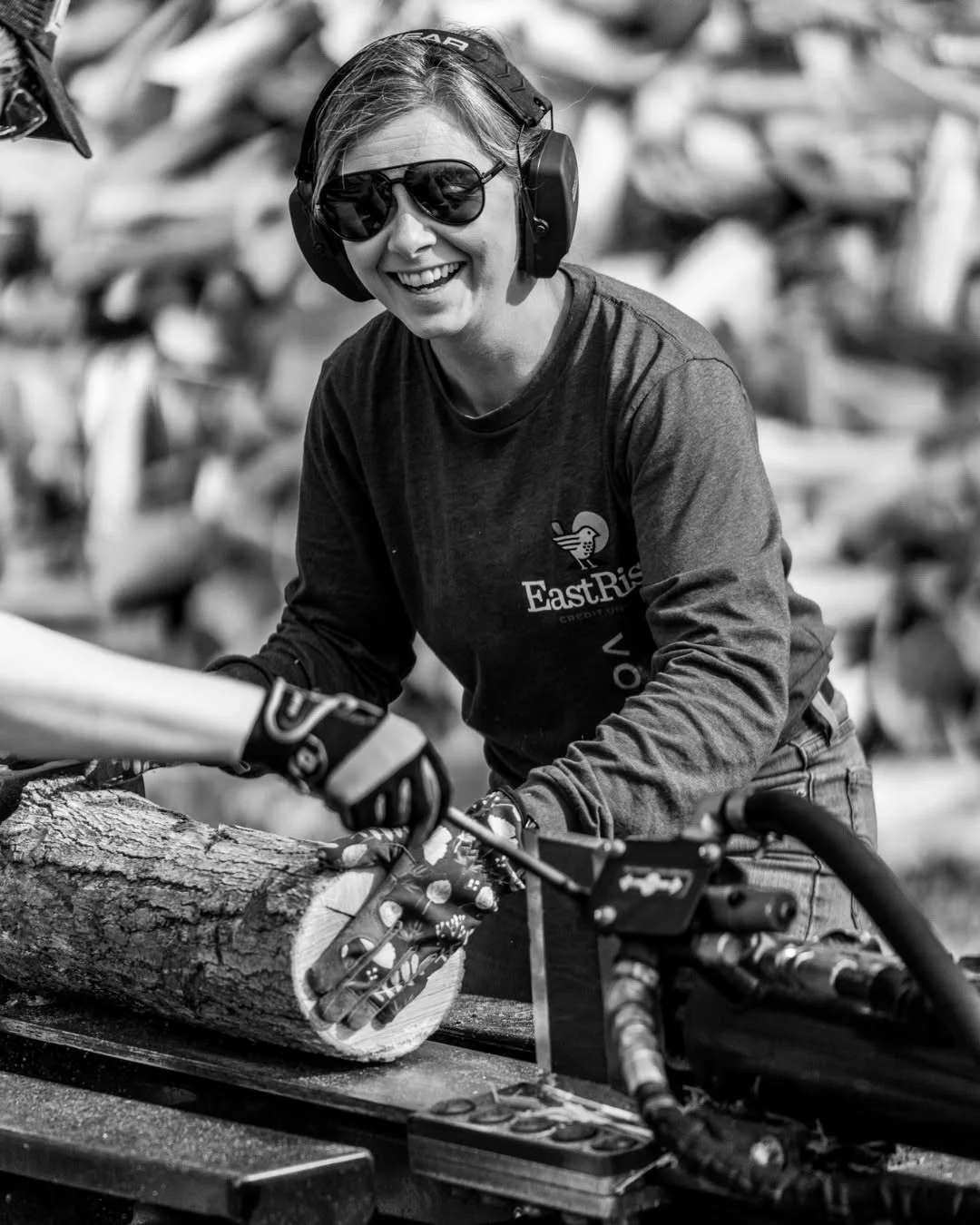 A smiling volunteer guides a log onto a splitter while wearing ear protection.