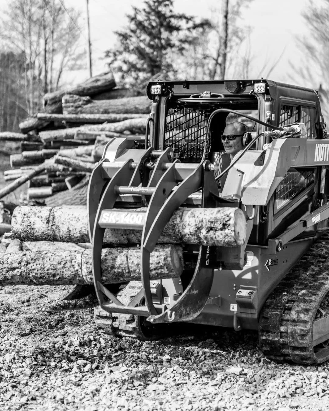 A tracked loader grips a bundle of logs beside stacked timber.