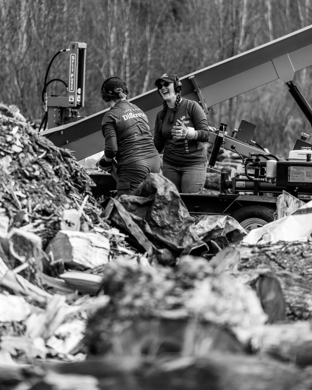 Two volunteers work beside a conveyor amid piles of split wood.