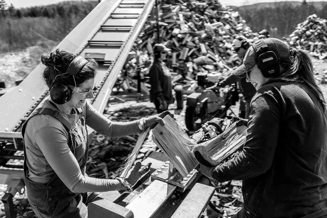 Two volunteers pass split logs across a conveyor beside a woodpile.