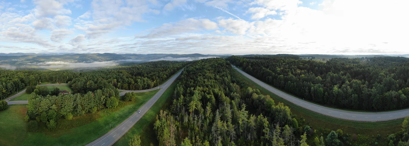 Panoramic aerial view of a divided highway curving through green forest beneath patterned clouds